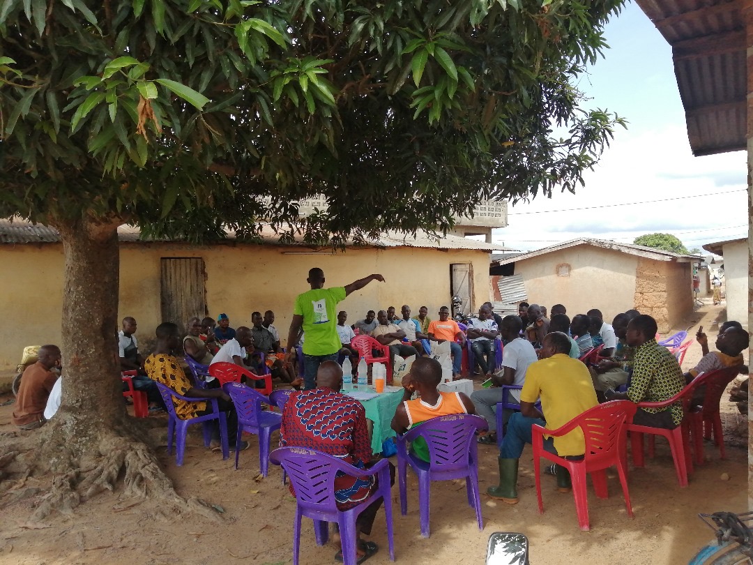 Réunion communautaire en plein air sous un arbre, animée par un intervenant en tee-shirt vert de l'entreprise AS Agri
